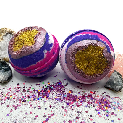 Two pink, purple and white bath bombs with a geode inspired center on a white surface with glitter scattered around.