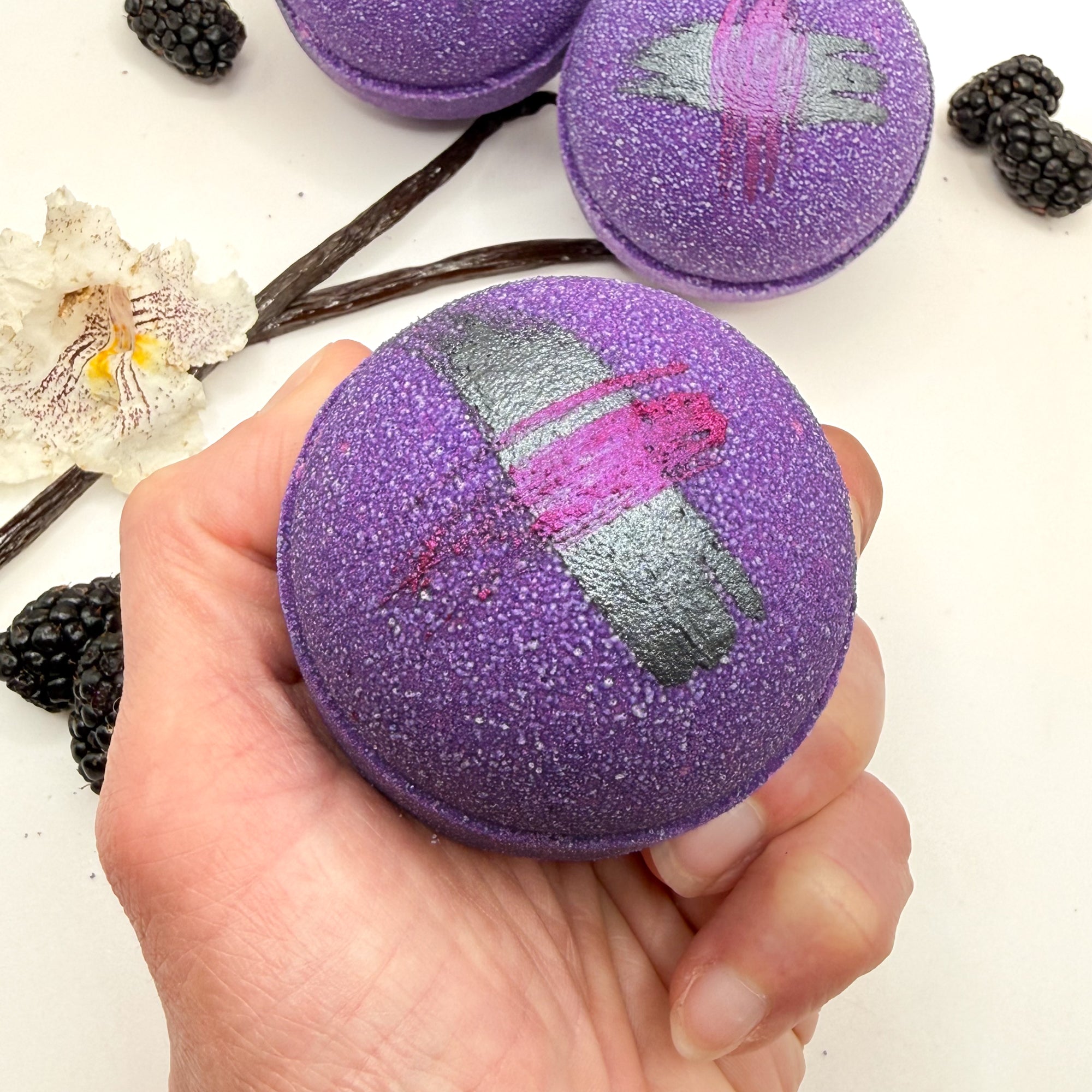Purple raspberry vanilla bath bomb held in a hand with blackberries and vanilla beans on a white background