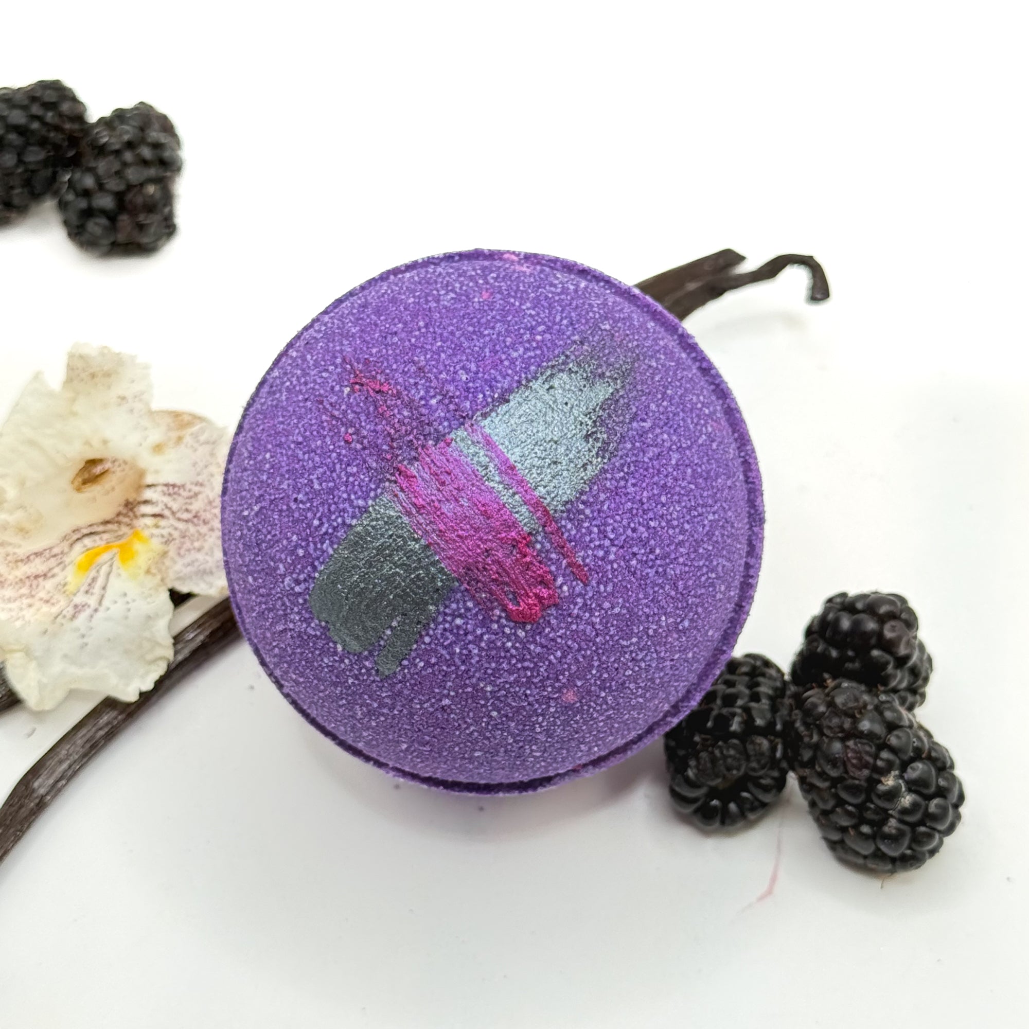 Purple raspberry vanilla bath bomb with a colorful design, surrounded by blackberries and vanilla beans on a white background.