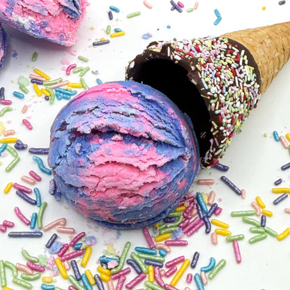 A scoop of lite purple and pink bubble bath scoop resembling a scoop of ice cream with a chocolate covered cone and rainbow sprinkles handmade by Rusted Acre Soap Co.