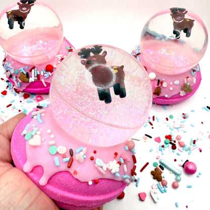 Pink donut-shaped bath bomb topped  with a reindeer bouncy ball to make a snow globe shape in a hand with colorful sprinkles on a white background. Handmade by Rusted Acre Soap Co.