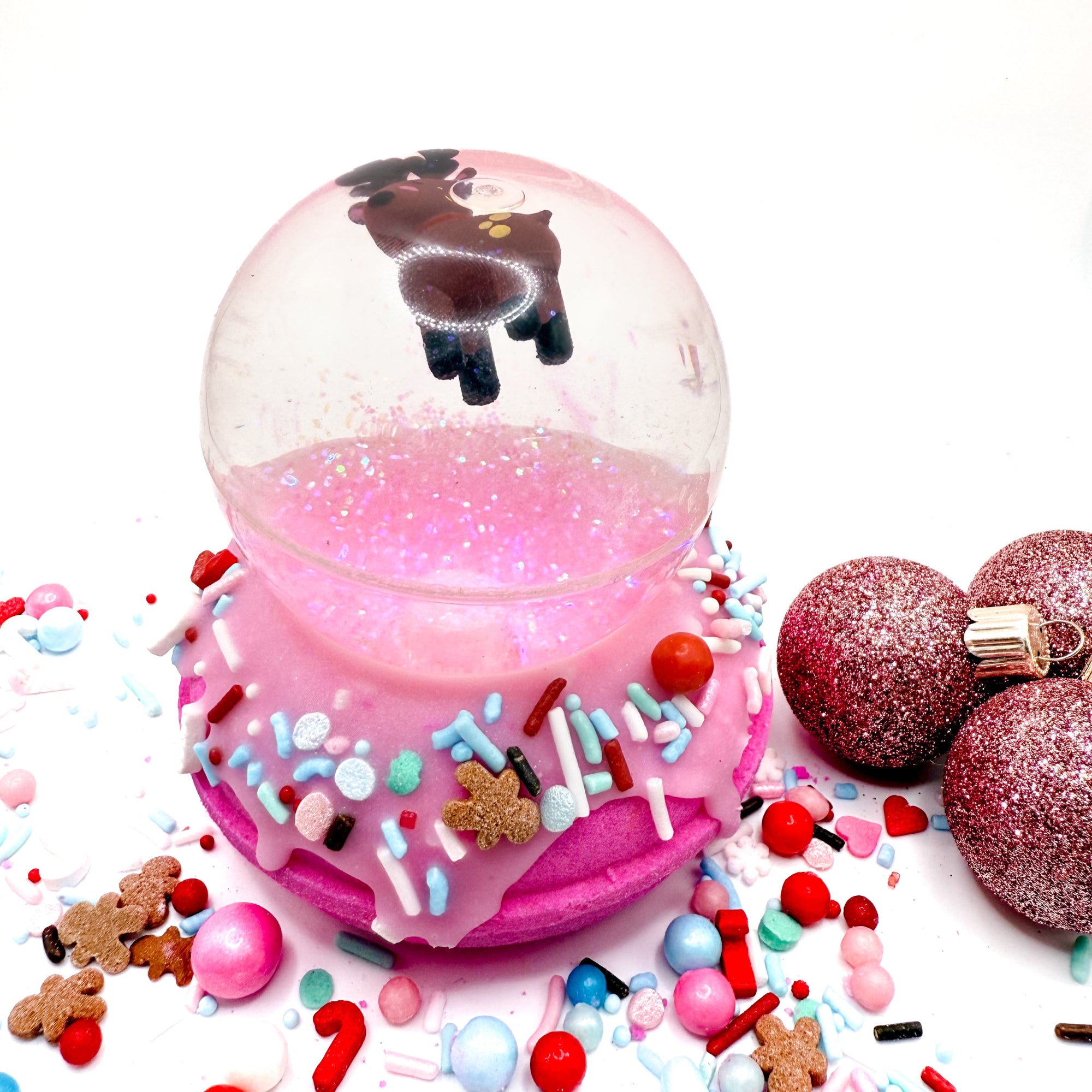 Pink donut bath bomb topped with a reindeer bouncy ball, surrounded by colorful sprinkles on a white background. Handmade by Rusted Acre Soap Co.