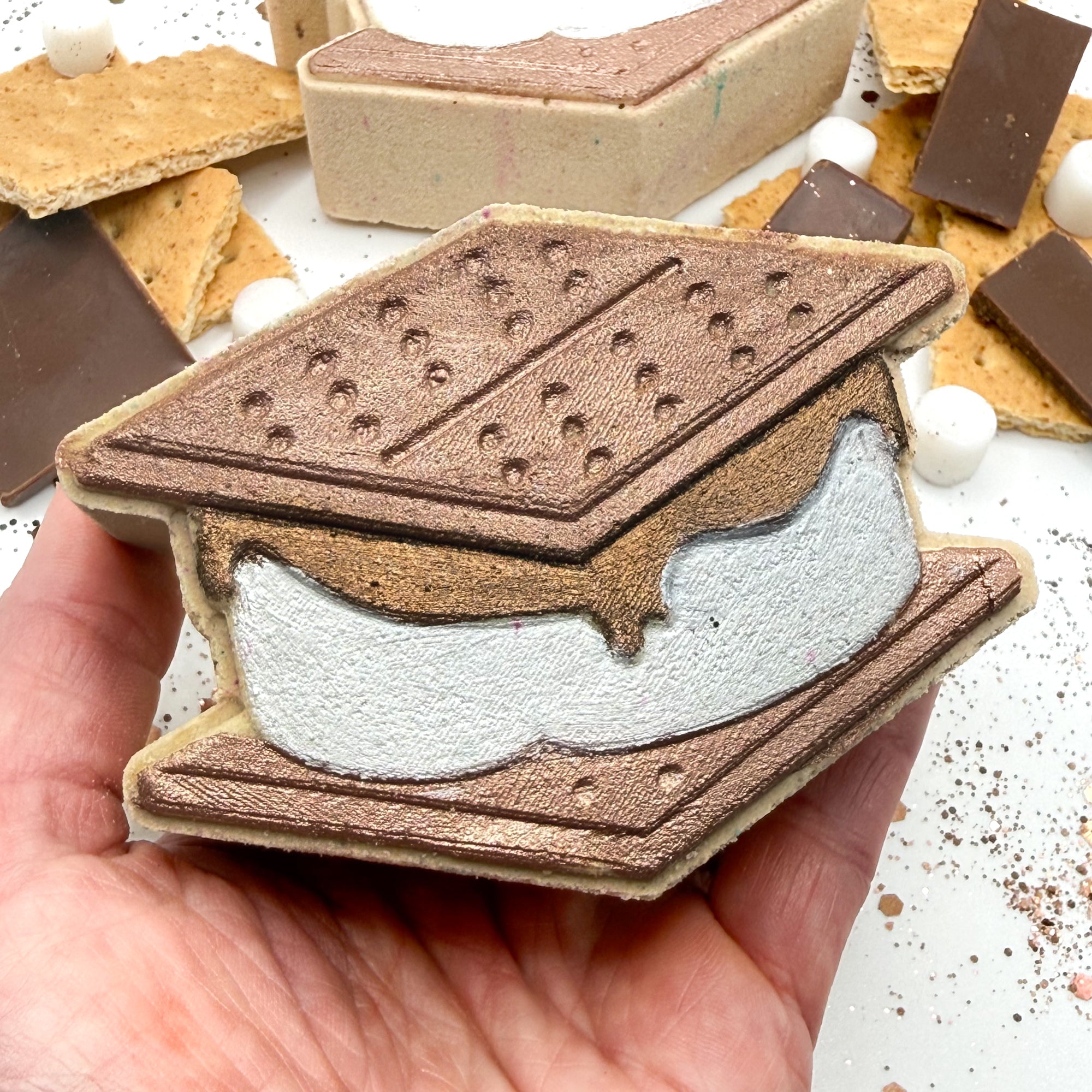 Hand holding a hand painted graham cracker and marshmallow stack bath bomb resembling a s'more.