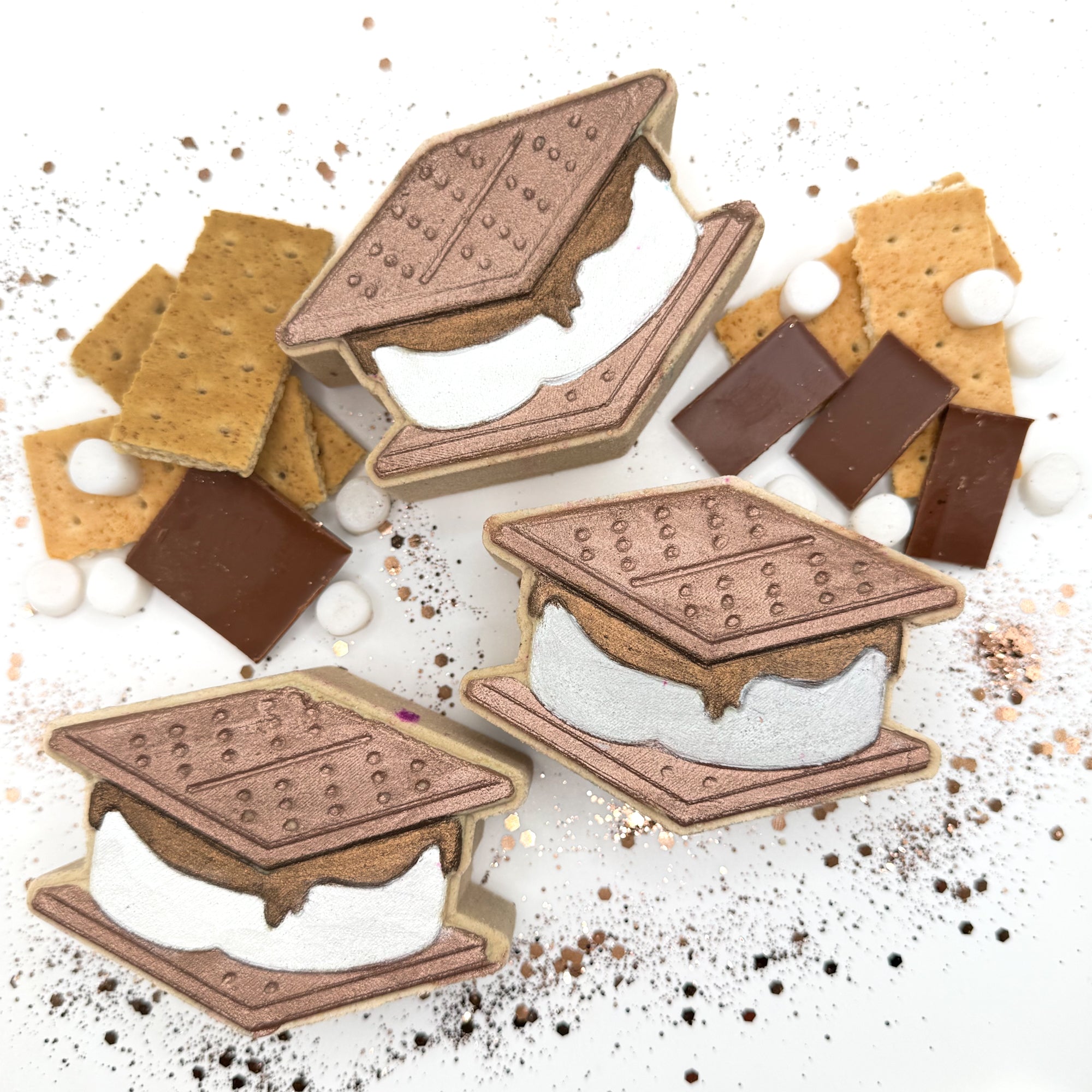 S'mores shaped bath bombs with graham crackers, chocolate, and marshmallows decorations on a white background.