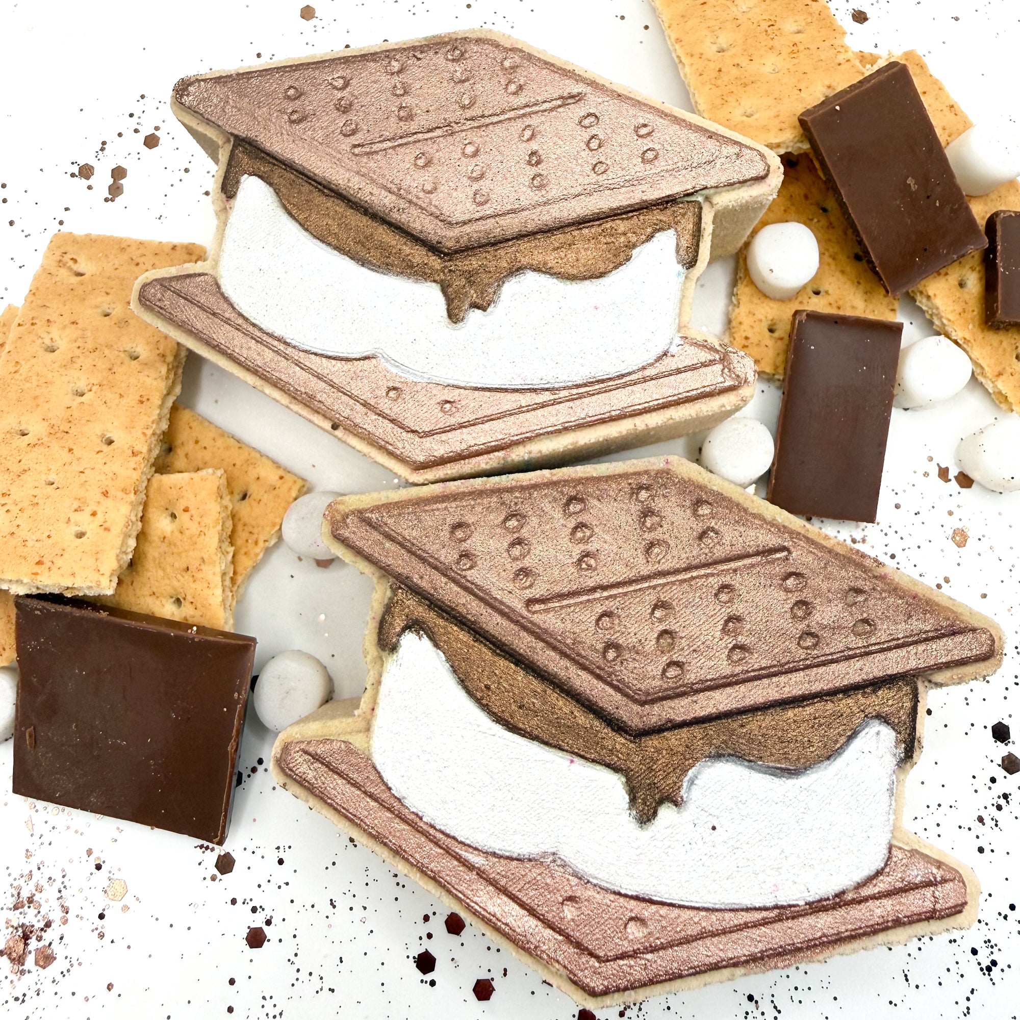 Handmade S'mores shaped bath bombs with graham crackers, chocolate, and marshmallows surrounding the bath bombs on a white background