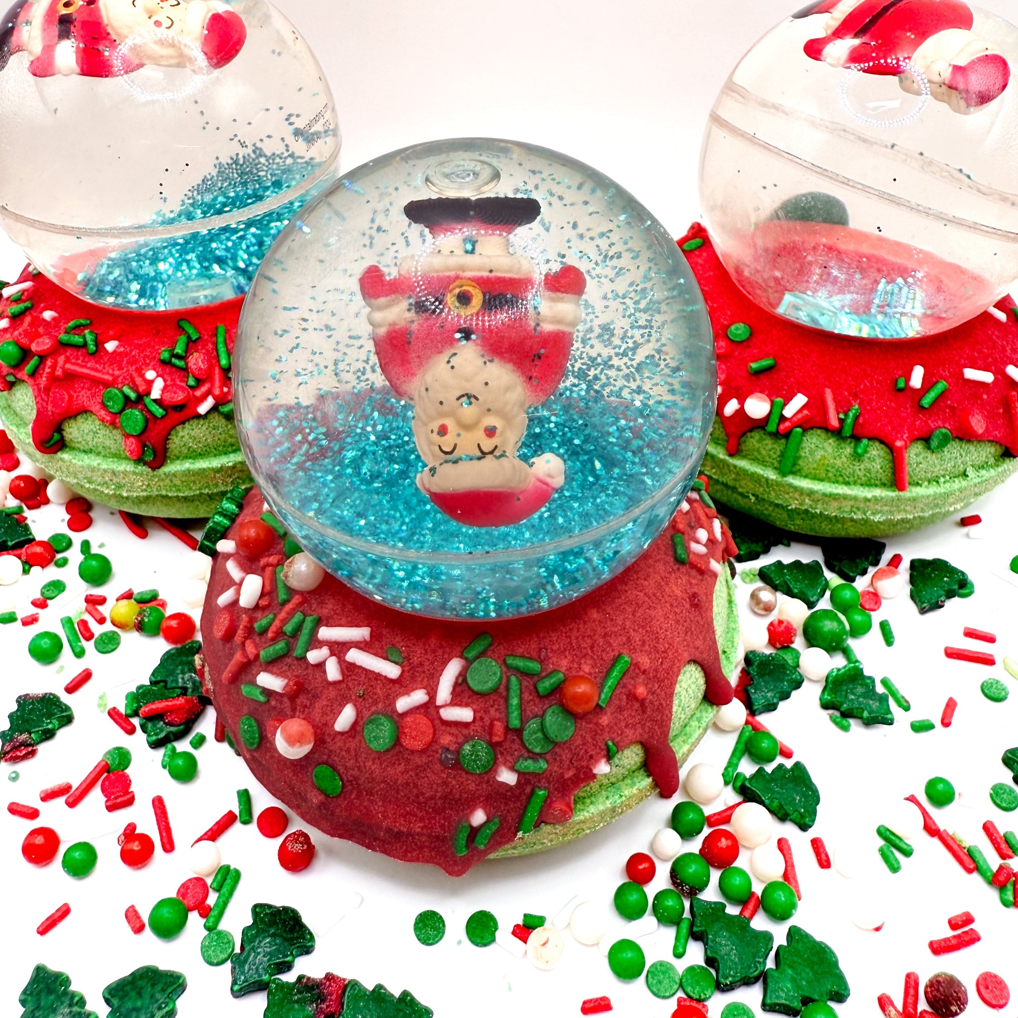 Three Santa snow globe bath bombs. Green donut shaped bath bombs with red glaze topped with a Santa bouncy balls. Surrounded with red and green festive sprinkles.