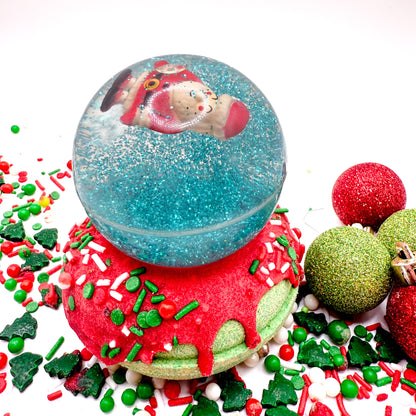A Santa snow globe bath bomb. Green donut shaped bath bomb with red glaze topped with a santa bouncy ball. Surrounded with red and green festive sprinkles and balls.
