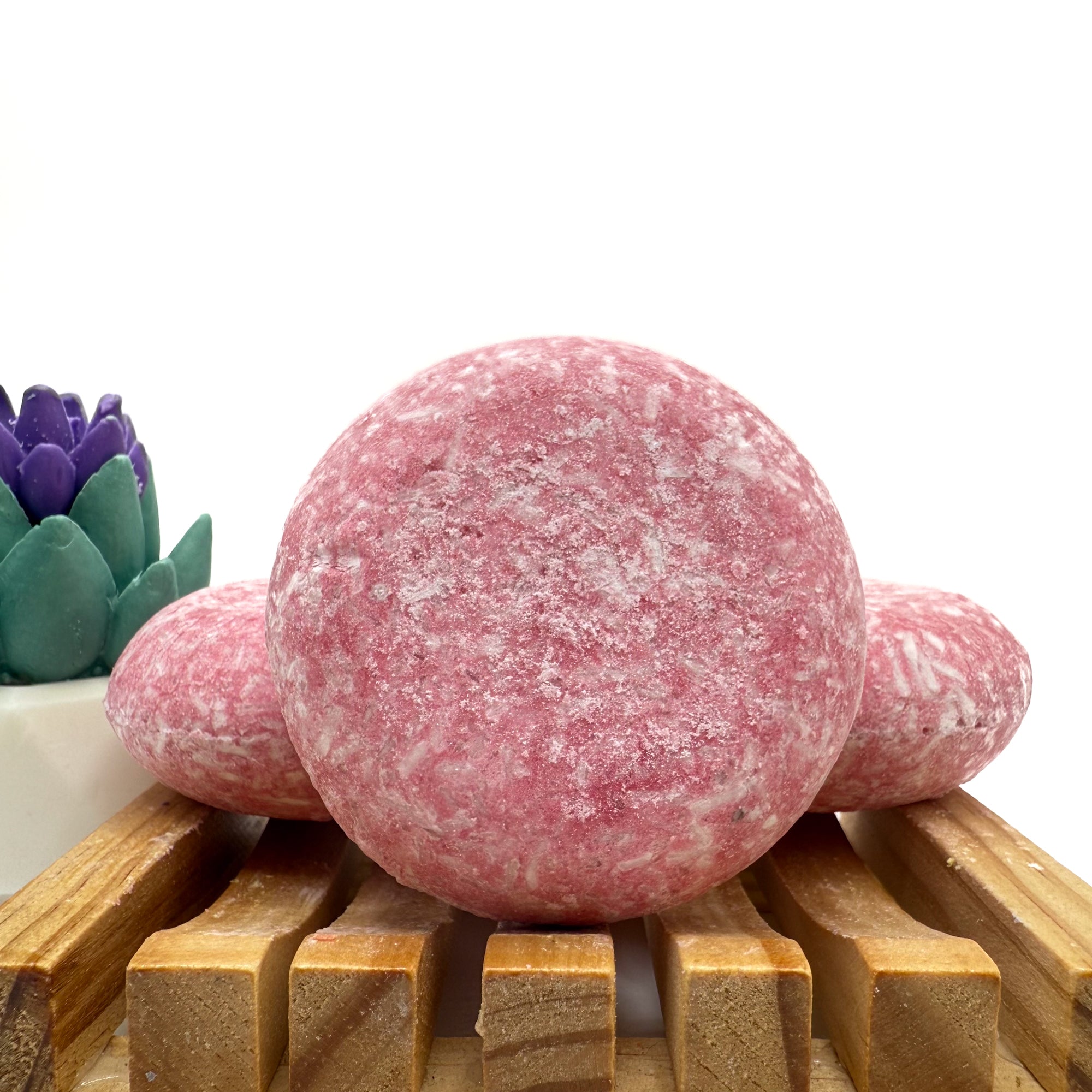 Three raspberry kiss shampoo bars sitting on a wood soap dish two laying flat, one standing up.