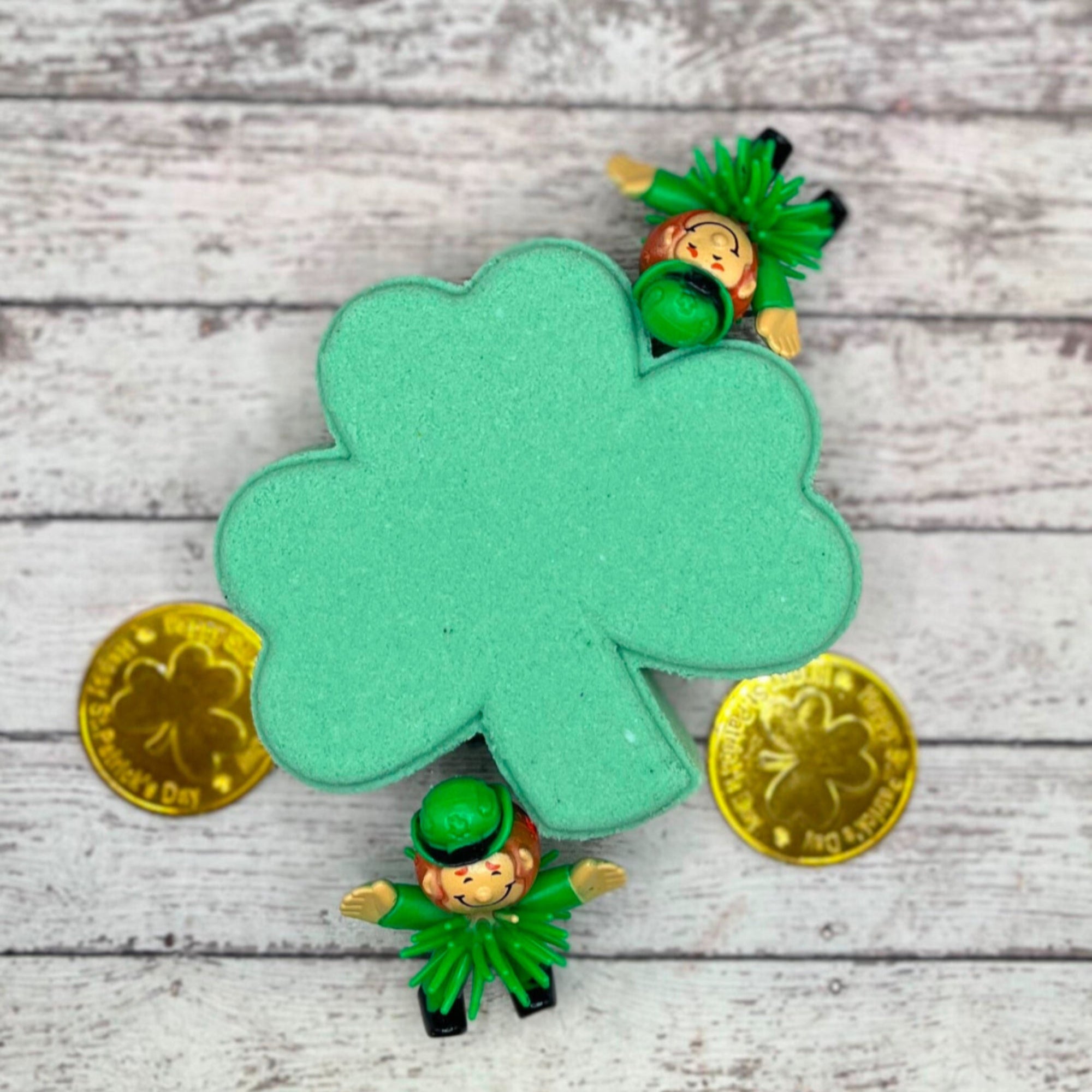 A green shamrock bath bomb handmade by Rusted Acre Soap Co. with surprise inside. Displayed with two leprechauns and two gold coins.