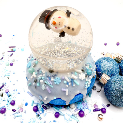 One snowman snow globe bath bomb hand made by Rusted Acre Soap Company. Bath bomb is a blue doughnut with white frosting and holiday sprinkles. Snowglobe is a bouncy ball with silver glitter photographed on a white background. 