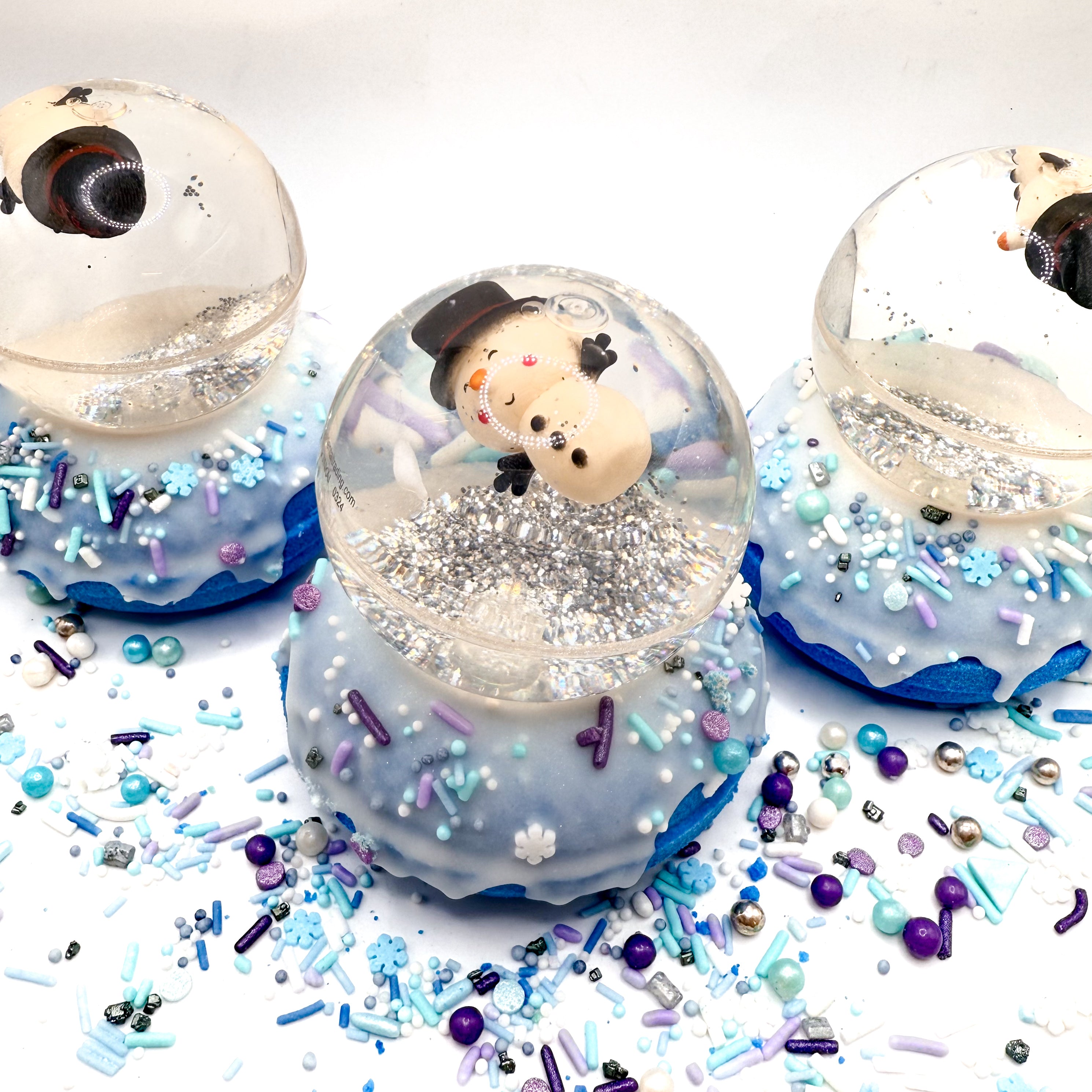 Three snowman snow globe bath bombs. Bath bomb is a blue doughnut with white frosting and holiday sprinkles. Snowglobe is a bouncy ball with silver glitter photographed on a white background. 