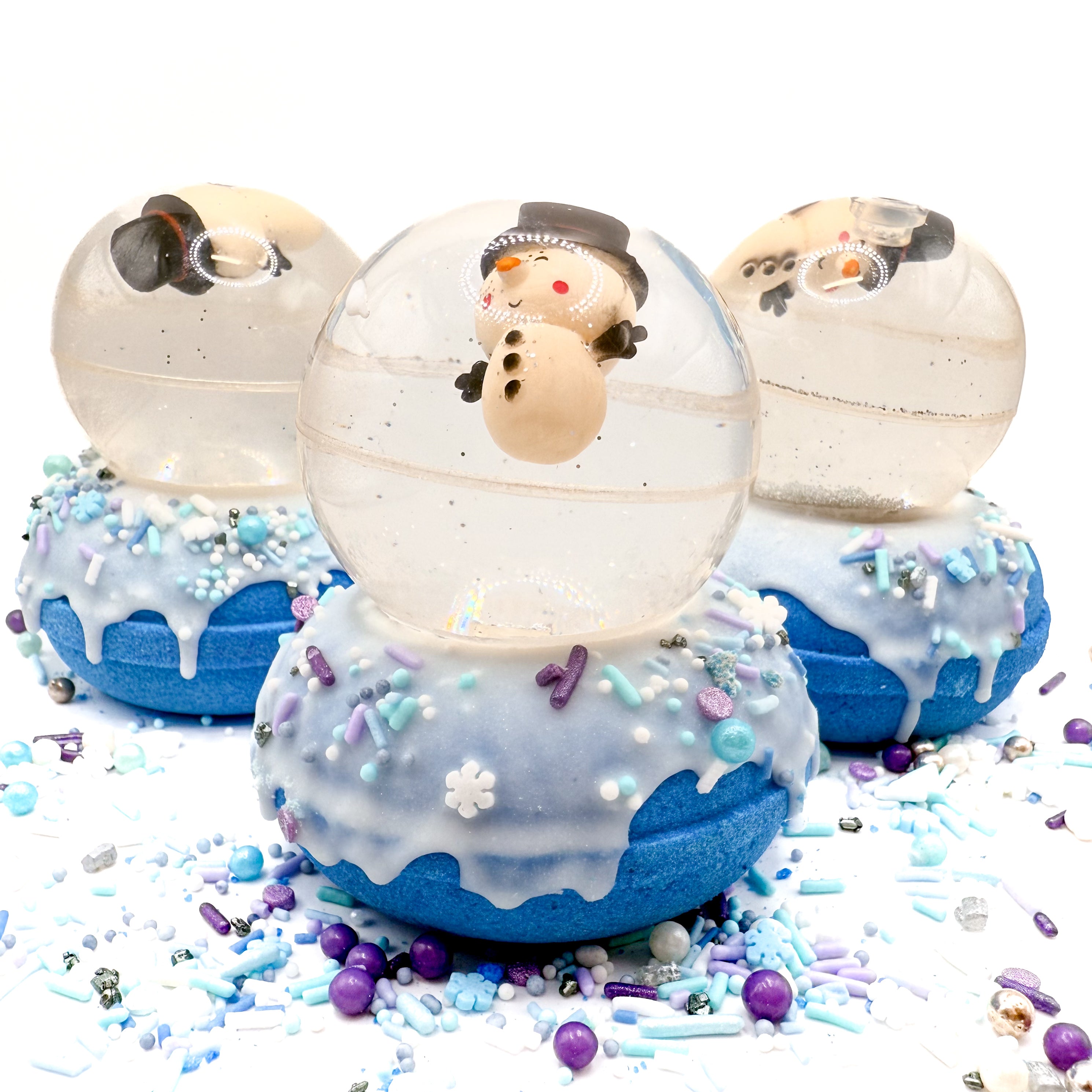 Three hand made snowman snow globe bath bombs sitting on a white background. Bath bomb is a blue doughnut with white frosting and holiday sprinkles. Snowglobe is a bouncy ball with silver glitter.