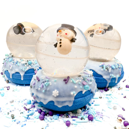 Three hand made snowman snow globe bath bombs sitting on a white background. Bath bomb is a blue doughnut with white frosting and holiday sprinkles. Snowglobe is a bouncy ball with silver glitter.