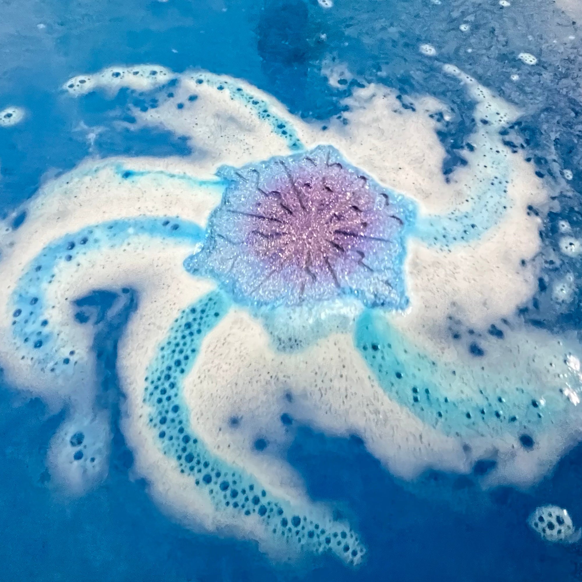 Foamy blue snowflake bath bomb in water  a pink and purple center. Made by Rusted Acre Soap Co.