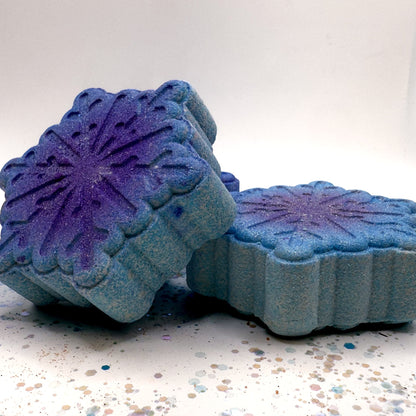 Two blue and purple snowflake shapped bath bombs on a speckled surface with a white background