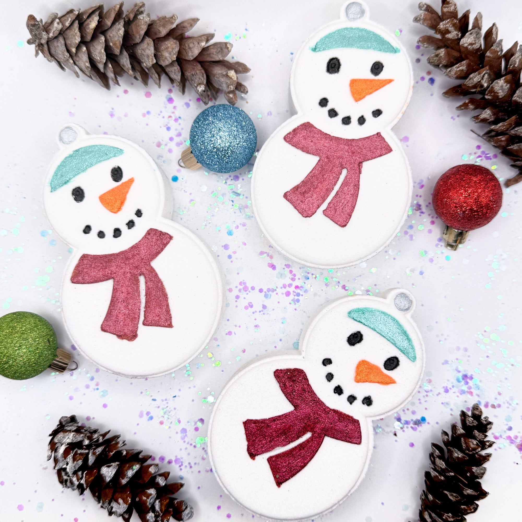 Three snowman bath bombs made by Rusted Acre Soap Co with red scarves on a white background with Christmas decorative elements.