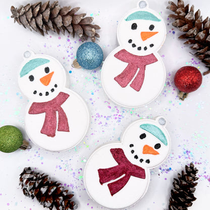 Three snowman bath bombs made by Rusted Acre Soap Co with red scarves on a white background with Christmas decorative elements.