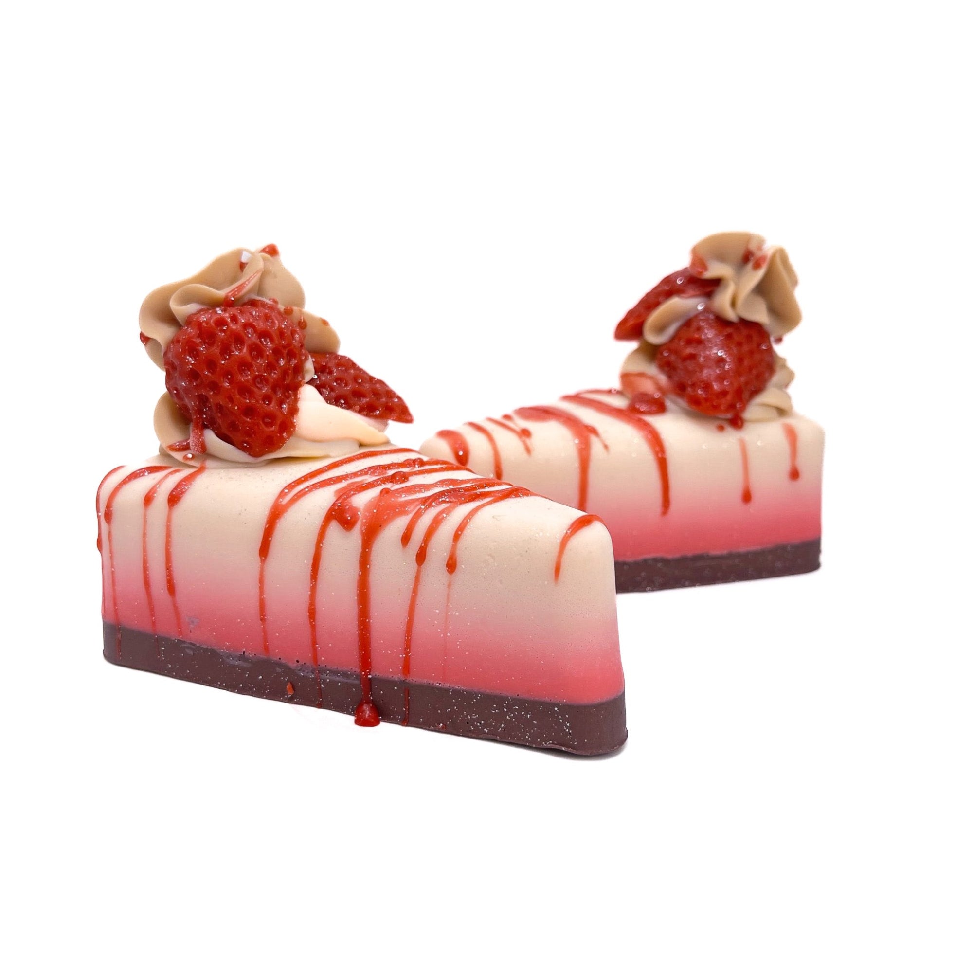 Two slices of strawberry cheesecake pie shape soap. Filling is white fading to red. The crust is dark. Topped with a cold process soap puff and red strawberry soap embeds. 