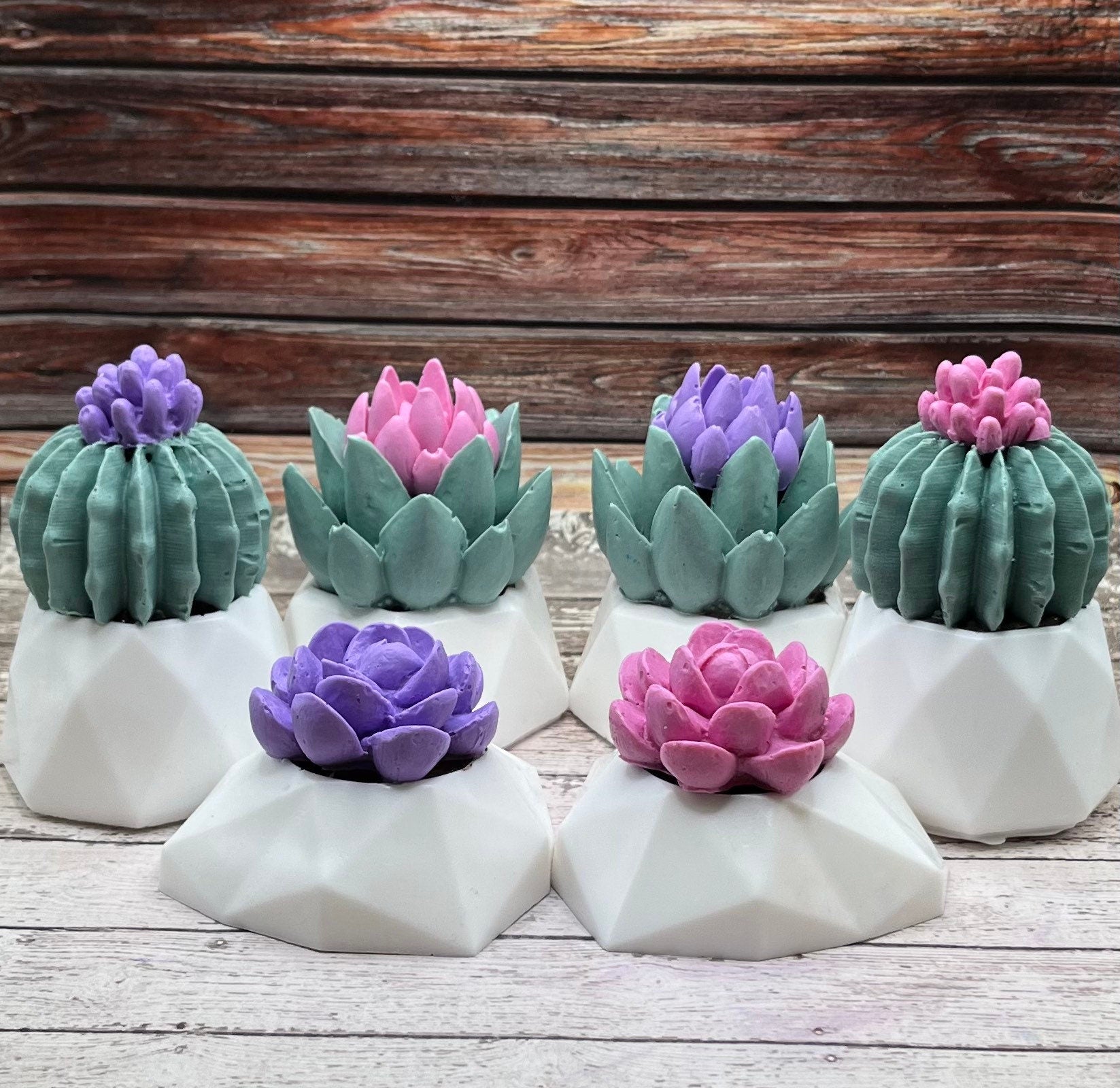 Various succulent shaped soaps displayed on a rustic wood background. Colors are pink, purple, and green with white pots. 