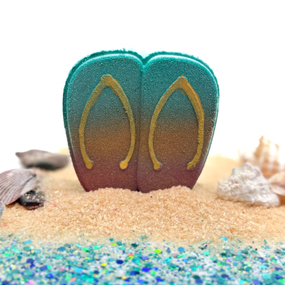 Hand painted teal and gold flip-flop-shaped bath bomb on a sandy surface with shells props and glitter.