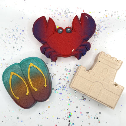 Three handmade beach themed bath bombs including flip-flops, a crab and a sandcastle on a sandy surface with shells and a sandcastle.