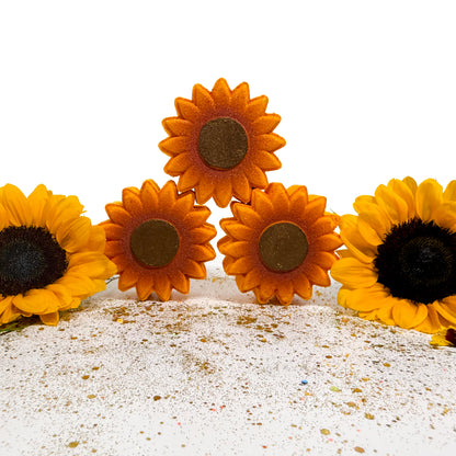 Four sunflower bath bombs arranged on a white surface with glittery white sand, with two flowers standing upright and two lying on their sides against a white background.