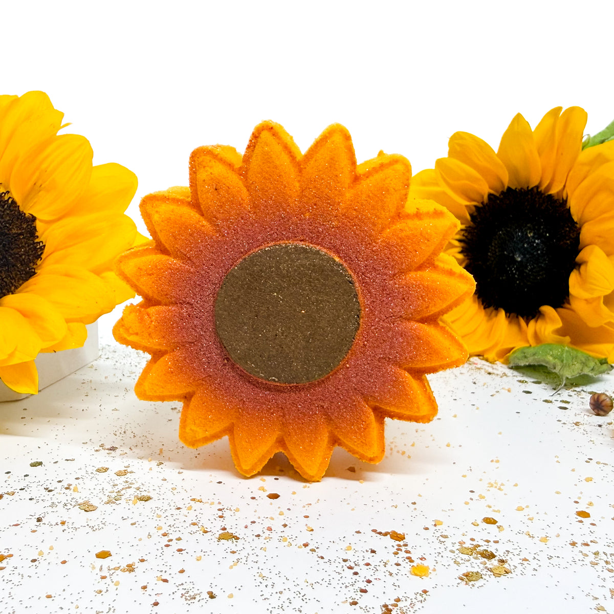 A sunflower-shaped bath bomb with orange and yellow petals, a dark brown center, and a glittery orange-brown base, displayed on a white surface with two larger sunflower props in the background.