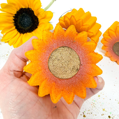 A hand holding a handmade sunflower-shaped bath bomb with orange petals, a glittery gold center, and a blurred yellow sunflower in the background.