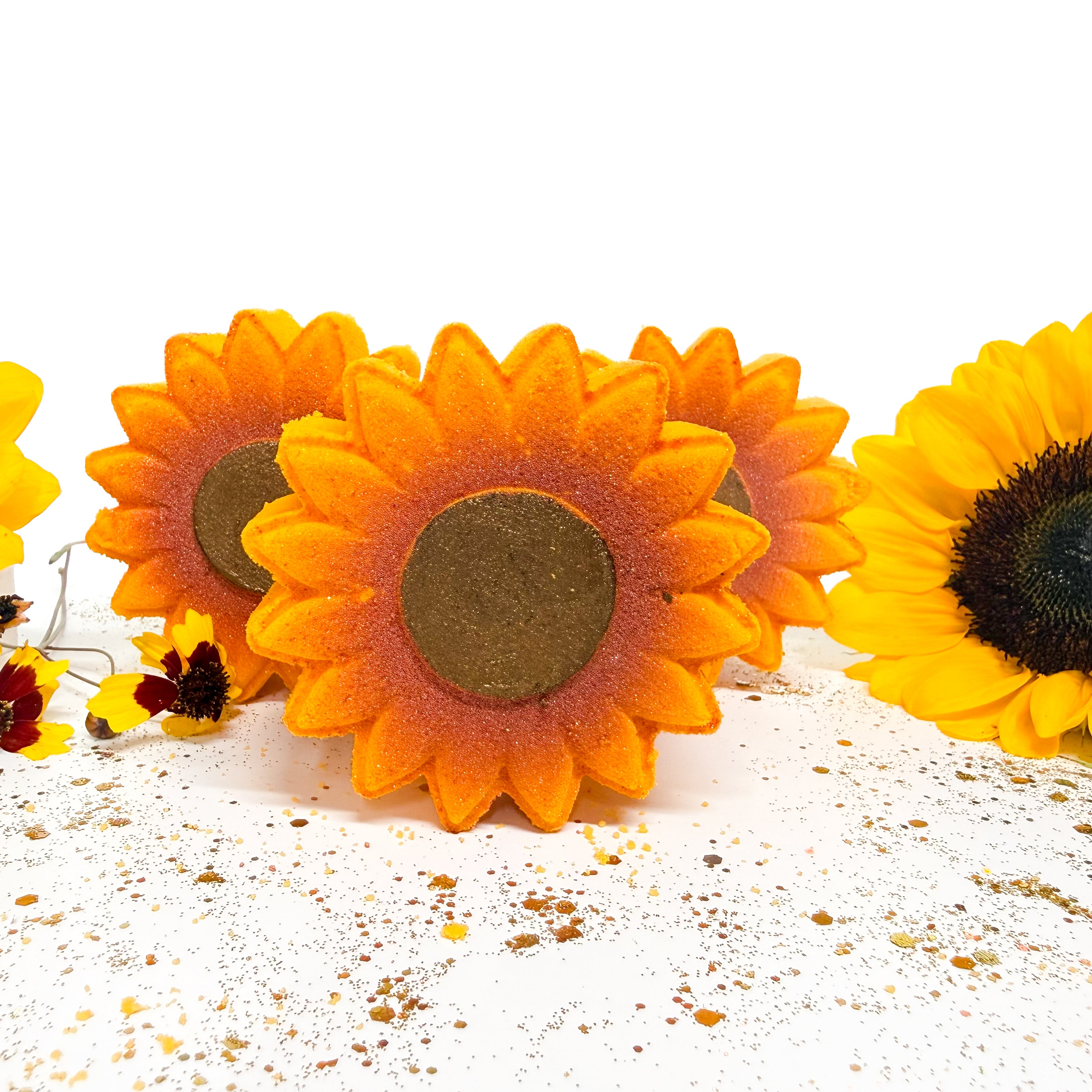 Three handmade sunflower bath bombswith orange and yellow petals and dark brown centers, arranged on a white surface with scattered flower petals.