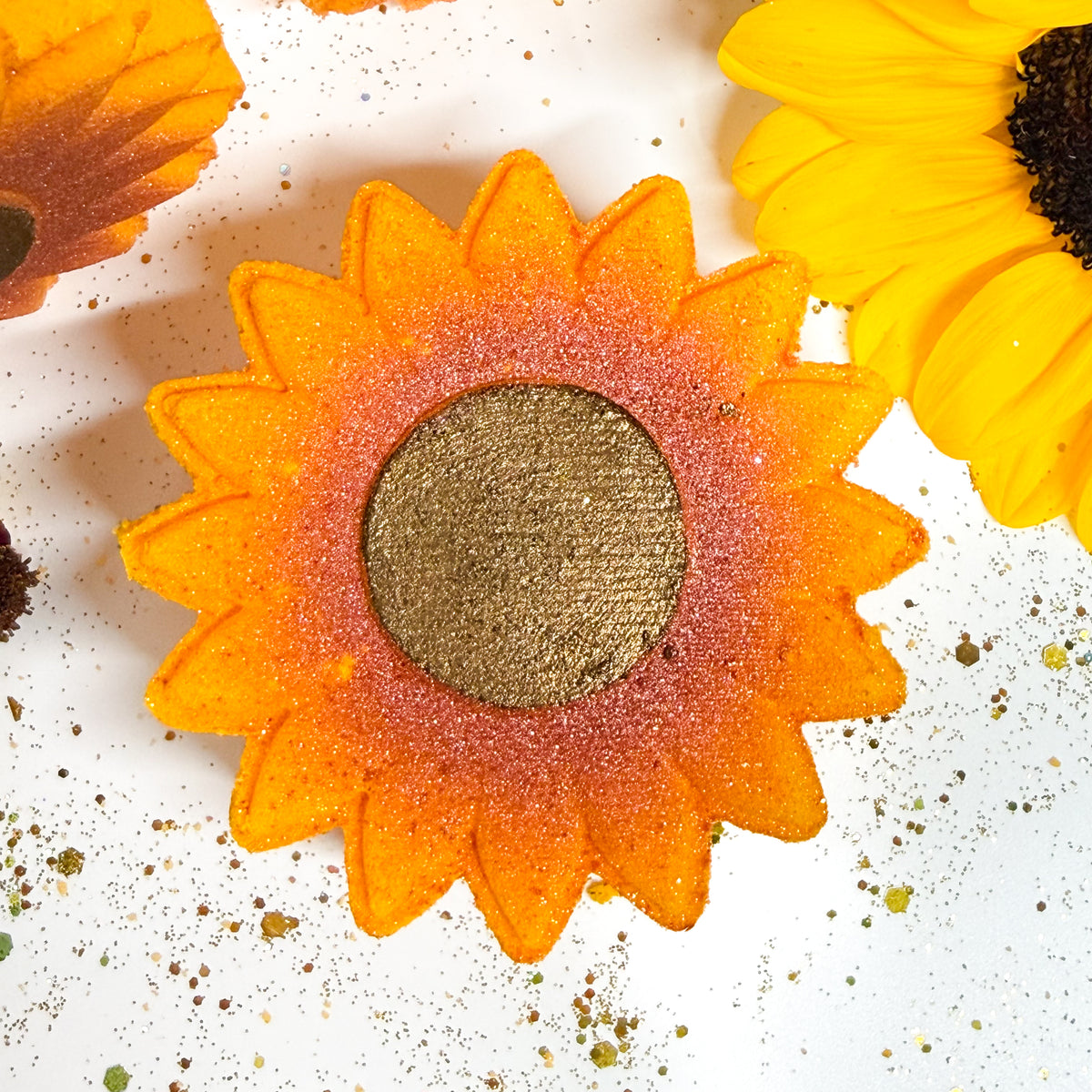A close-up of a sunflower-shaped bath bomb, likely an eyeshadow or face paint palette, with orange and yellow petals and a textured gold center, surrounded by loose gold glitter on a white background with partial sunflower props.
