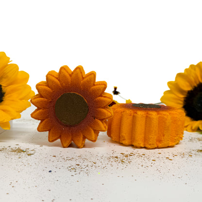 Two handmade sunflower-shaped bath bombs on a white surface with scattered glitter, flanked by partial yellow sunflowers against a white background.