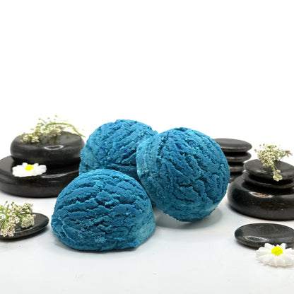 Three handmade blue bubble bath scoops that look like ice cream with spa stones and loose botanicals for a backdrop. 