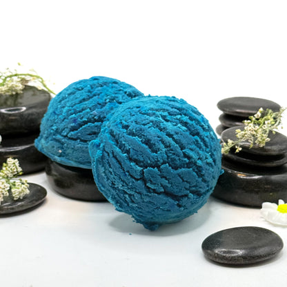 Two blue bubble bath scoops that look like ice cream with spa stones and loose botanicals for a backdrop. 