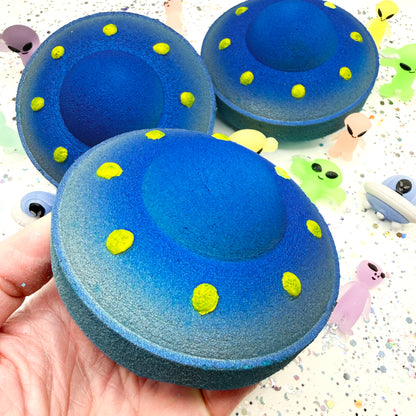 Blue UFO shaped bath bomb with yellow dots held in a hand against a colorful background.