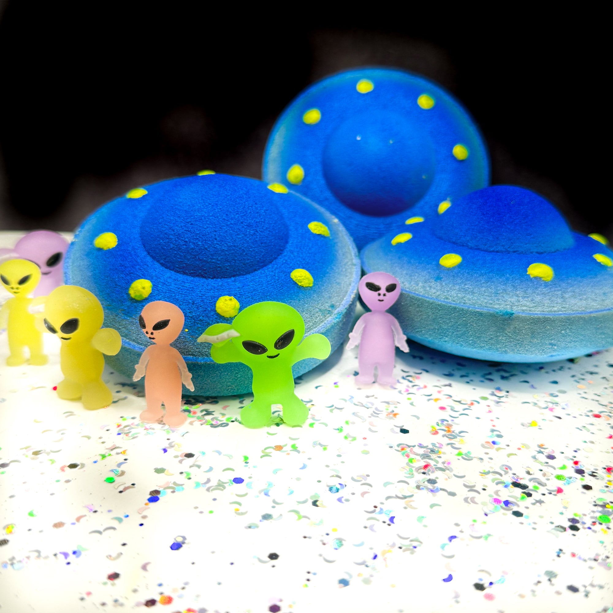 Three blue and yellow UFO bath bombs on a white background with alien toys placed around the bath bombs.