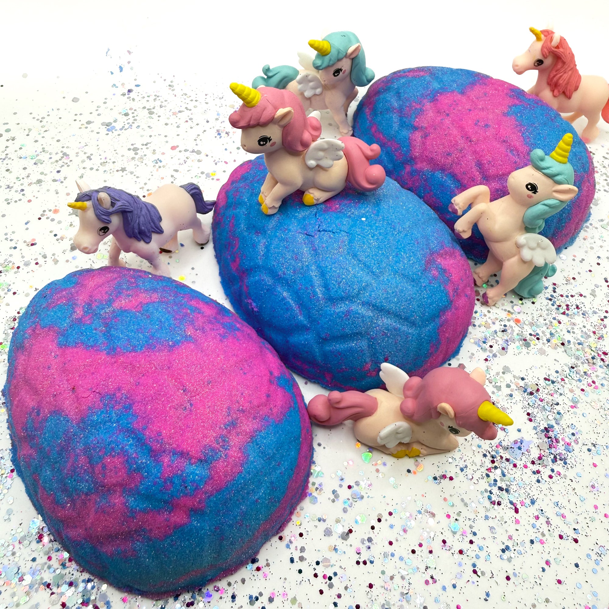 Three blue and pink unicorn egg bath bombs with unicorn figurines on a glittery surface.