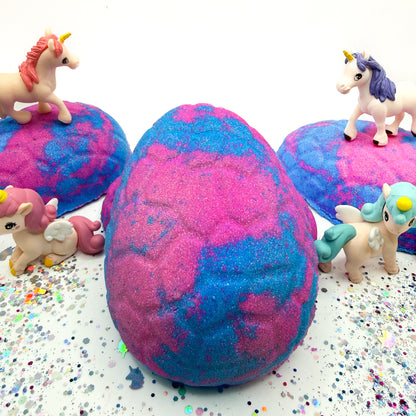 Handmade pink and blue unicorn egg bath bombs with unicorn figurines on a glittery white background.