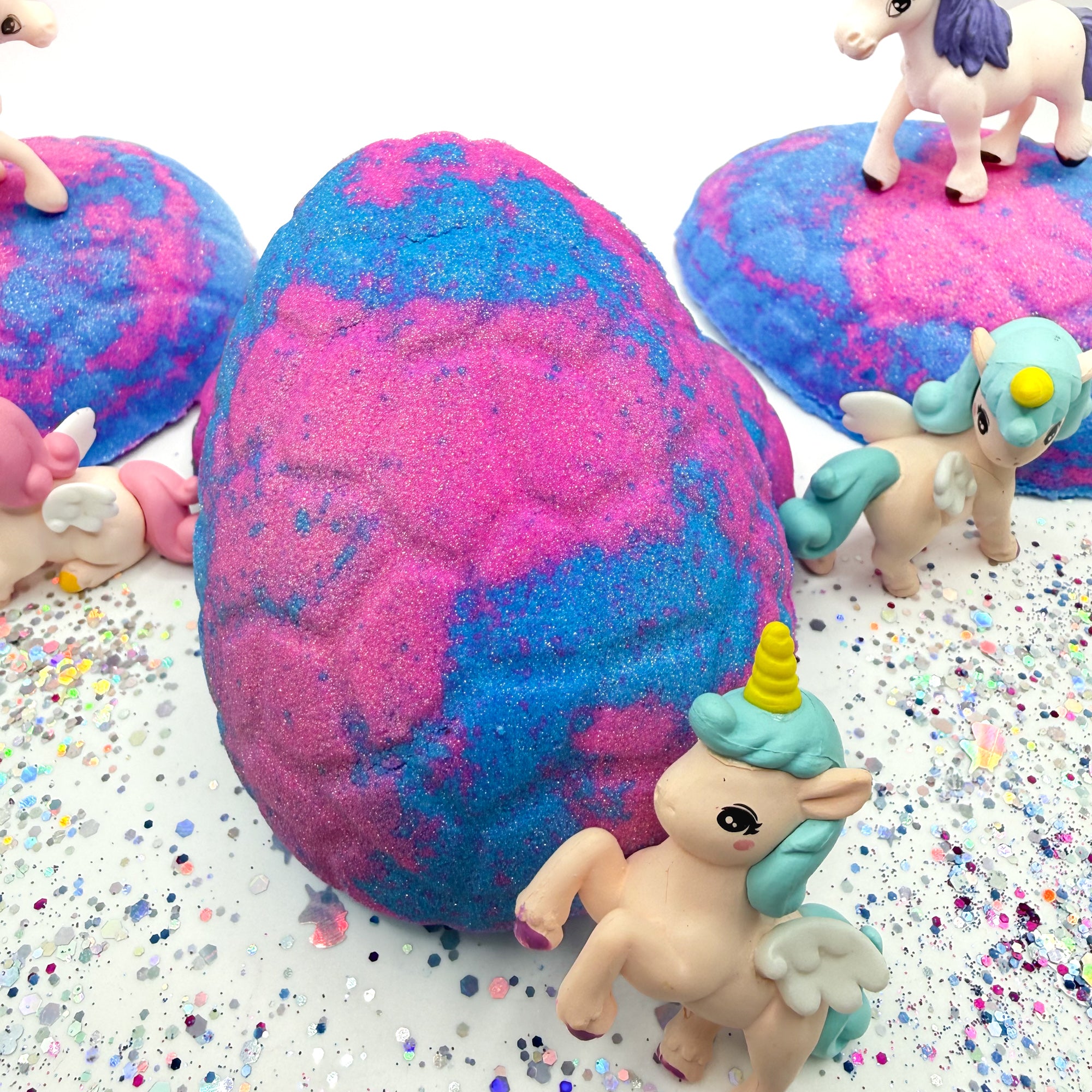 Handmade pink and blue unicorn egg bath bombs with unicorn figurines on a glittery surface. 