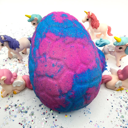 Unicorn egg bath bomb in pink and blue colors with unicorn figurines surrounding the bath bomb on a white background. 