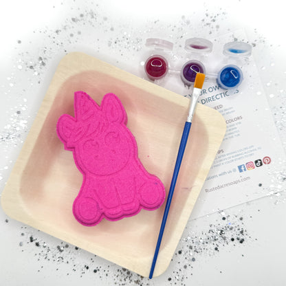 Full kit view of one pink paint your own unicorn shaped bath bomb displayed with a paint brush, paint pots with color.