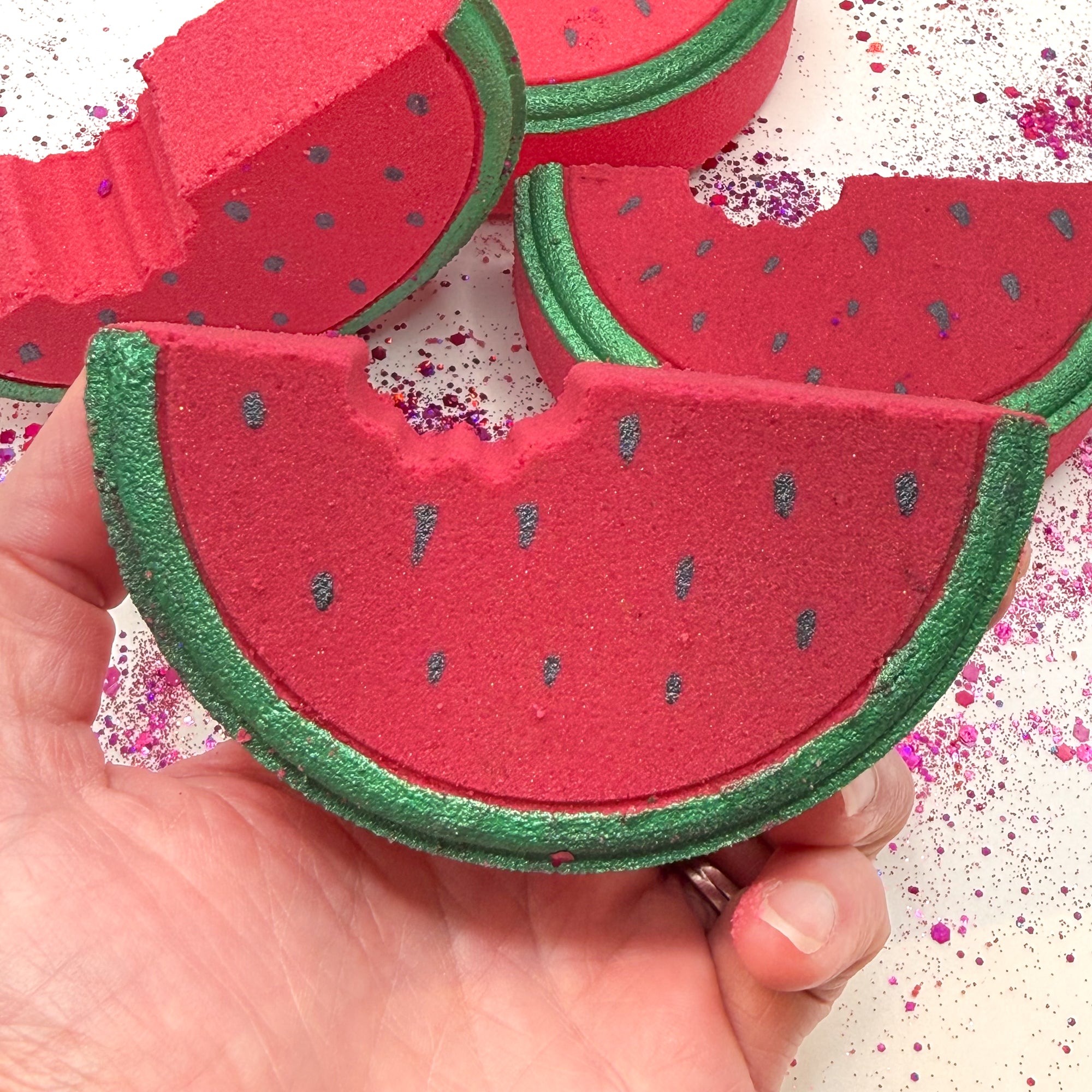 Up-close image of a red and green watermelon bath bomb with hand painted details being held in a hand with three other watermelon bath bombs in the background