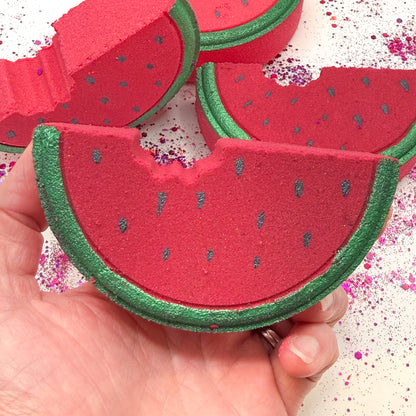Up-close image of a red and green watermelon bath bomb with hand painted details being held in a hand with three other watermelon bath bombs in the background