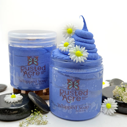 Two jars of blue tranquility whipped soap made by Rusted Acre Soap Co with spa type props in the background. 