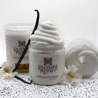 Three jars of vanilla whipped soap  made by Rusted Acre Soap Co surrounded by vanilla beans and flowers on a white background.