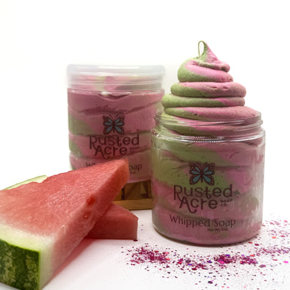 Two jars of Rusted Acre pink and green swirled watermelon twist whipped soap with a slice of watermelon on a white background
