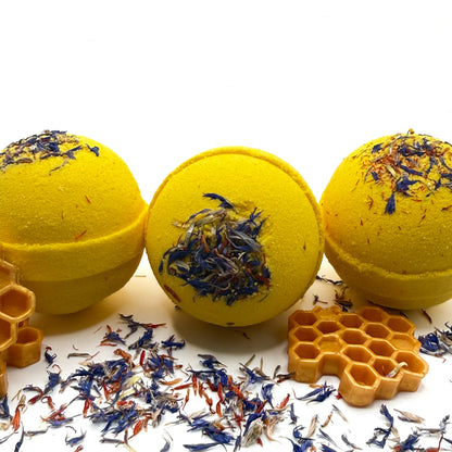 Three yellow wildflower honey bath bombs with floral wildflower  elements on a white background, surrounded by wildflower petals and a honeycomb soap.