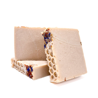 Three bars of handmade natural colored wildflower and honey soap from Rusted Acre Soap Co. Two upright and one lying flat to show texture. Red and blue flower petals on top. 