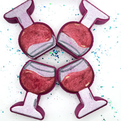 Four hand painted burgundy wine glass shaped bath bombs on a white background with blue and green glitter. Hand made by Rusted Acre Soap Co.