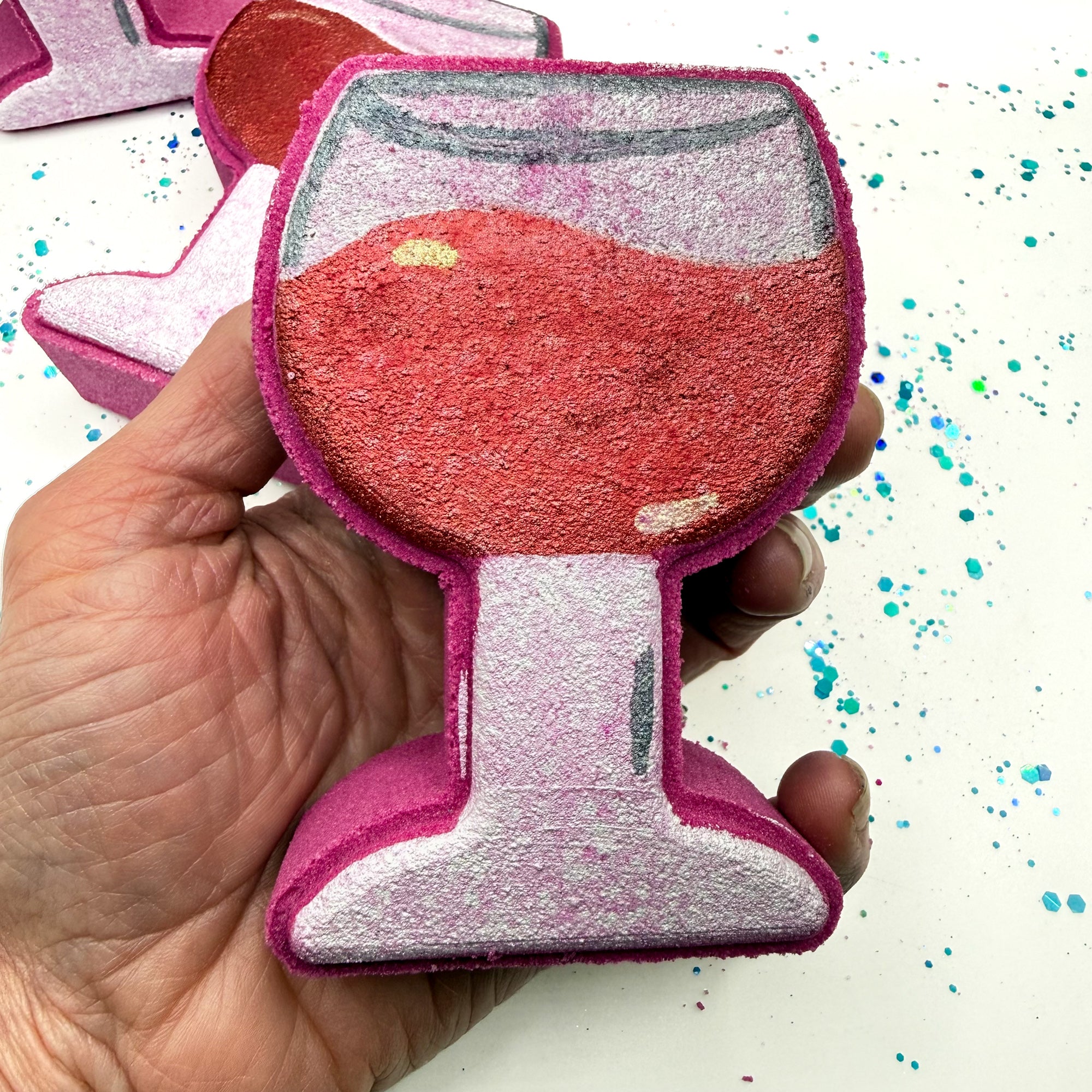One hand painted burgundy wine glass shaped bath bomb held in a hand with a white background with blue and green glitter. Made by Rusted Acre Soap Co.