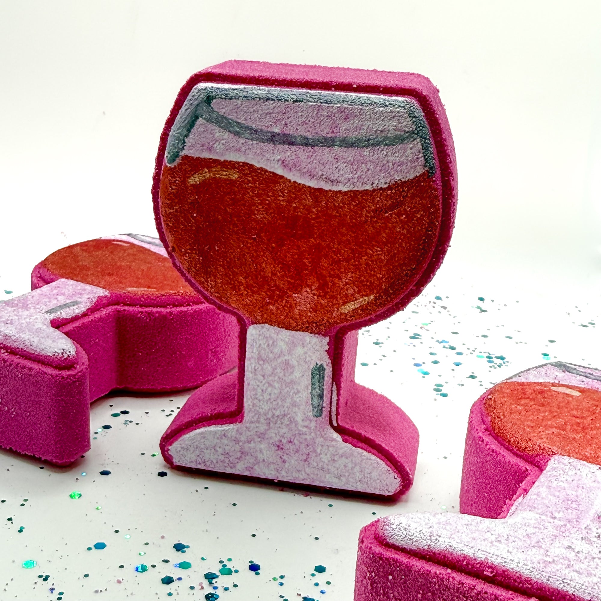 Three hand painted burgundy wine glass shaped bath bombs on a white background with blue and green glitter. Made by Rusted Acre Soap Co.